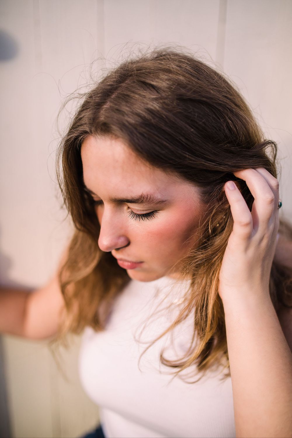 Young woman in white top with eyes closed touching her brown hair against a light background.