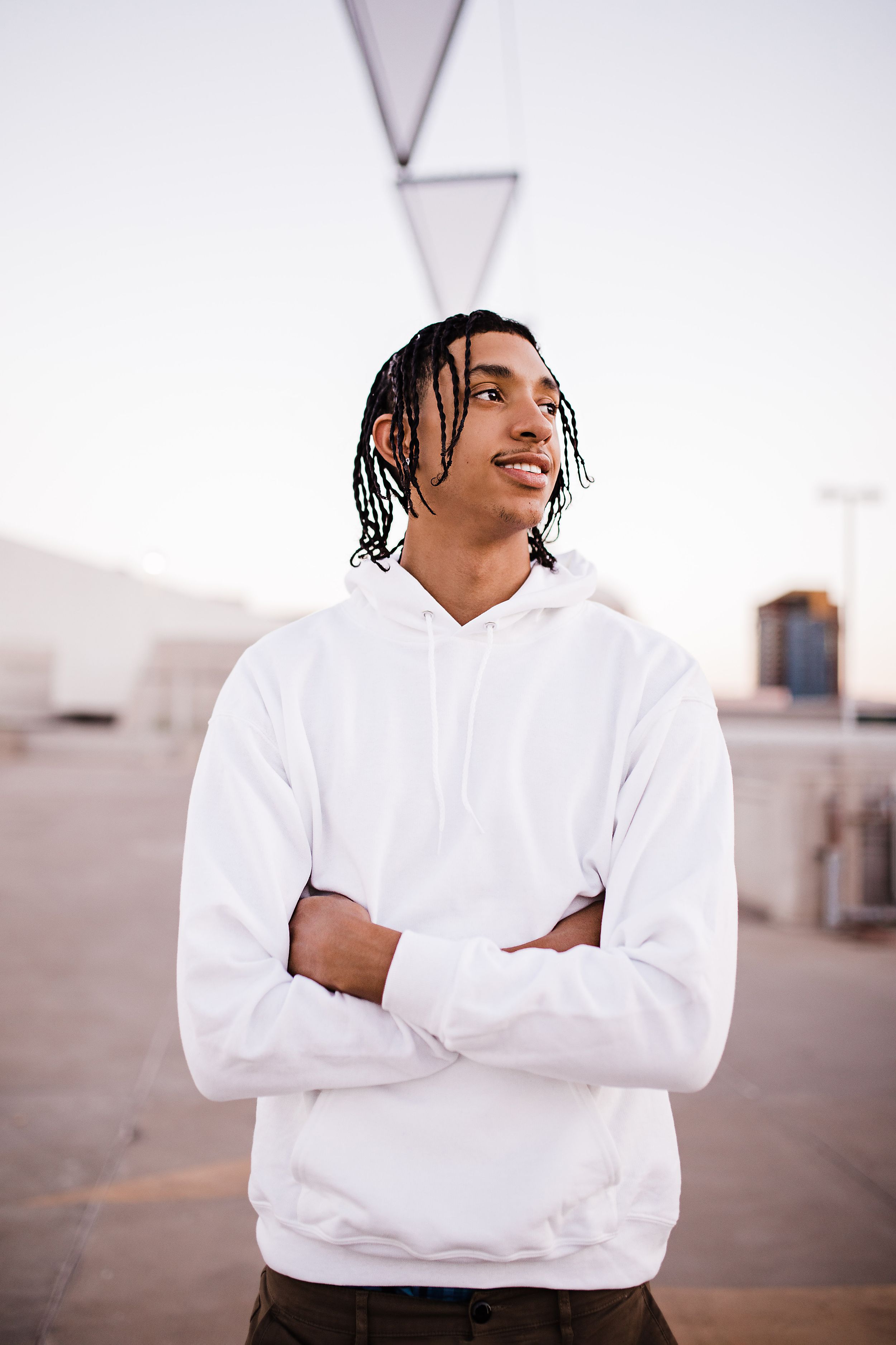 Young man with braids in white hoodie standing with arms crossed outdoors at sunset.