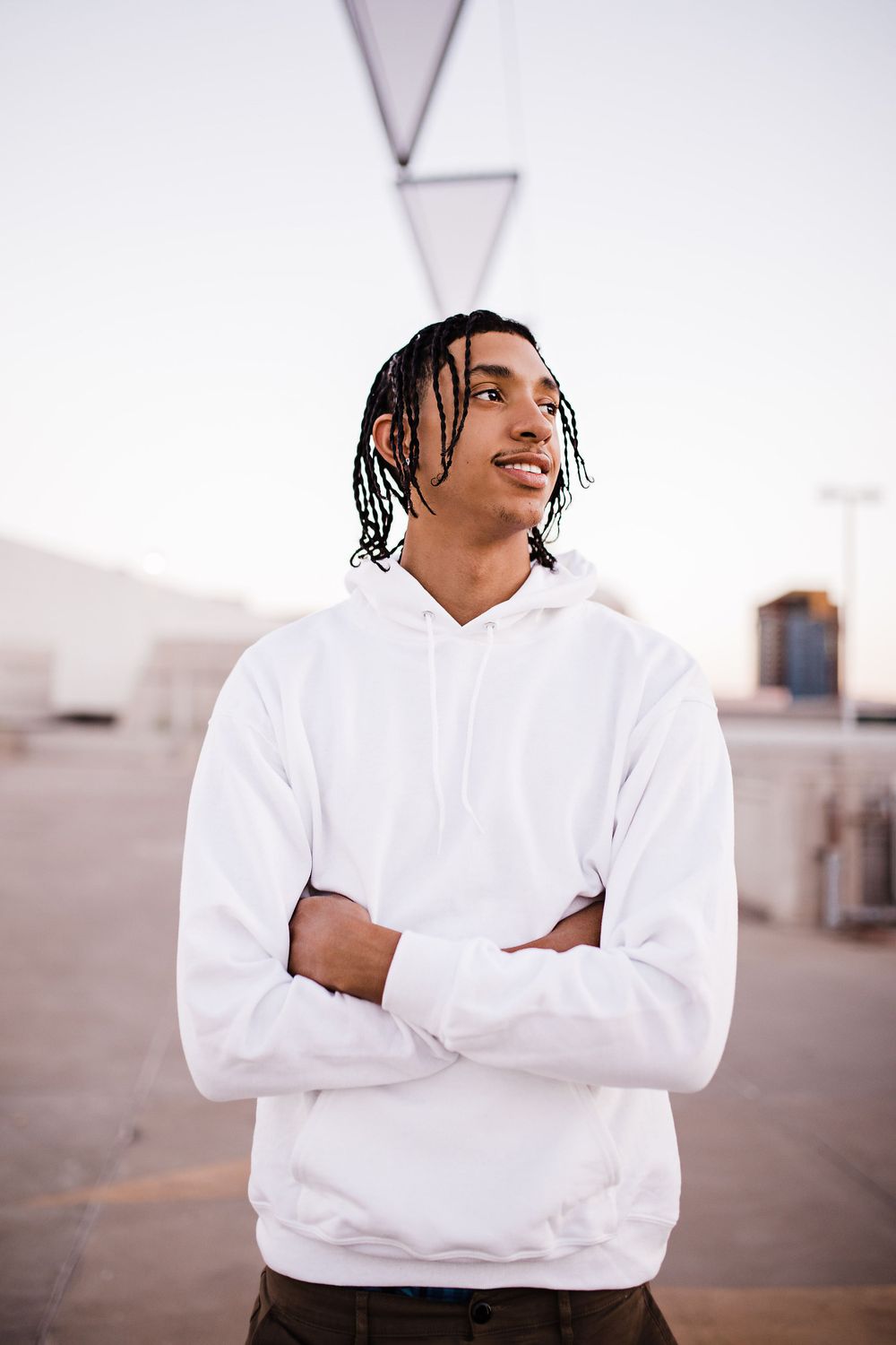 Young man with braids in white hoodie standing with arms crossed outdoors at sunset.