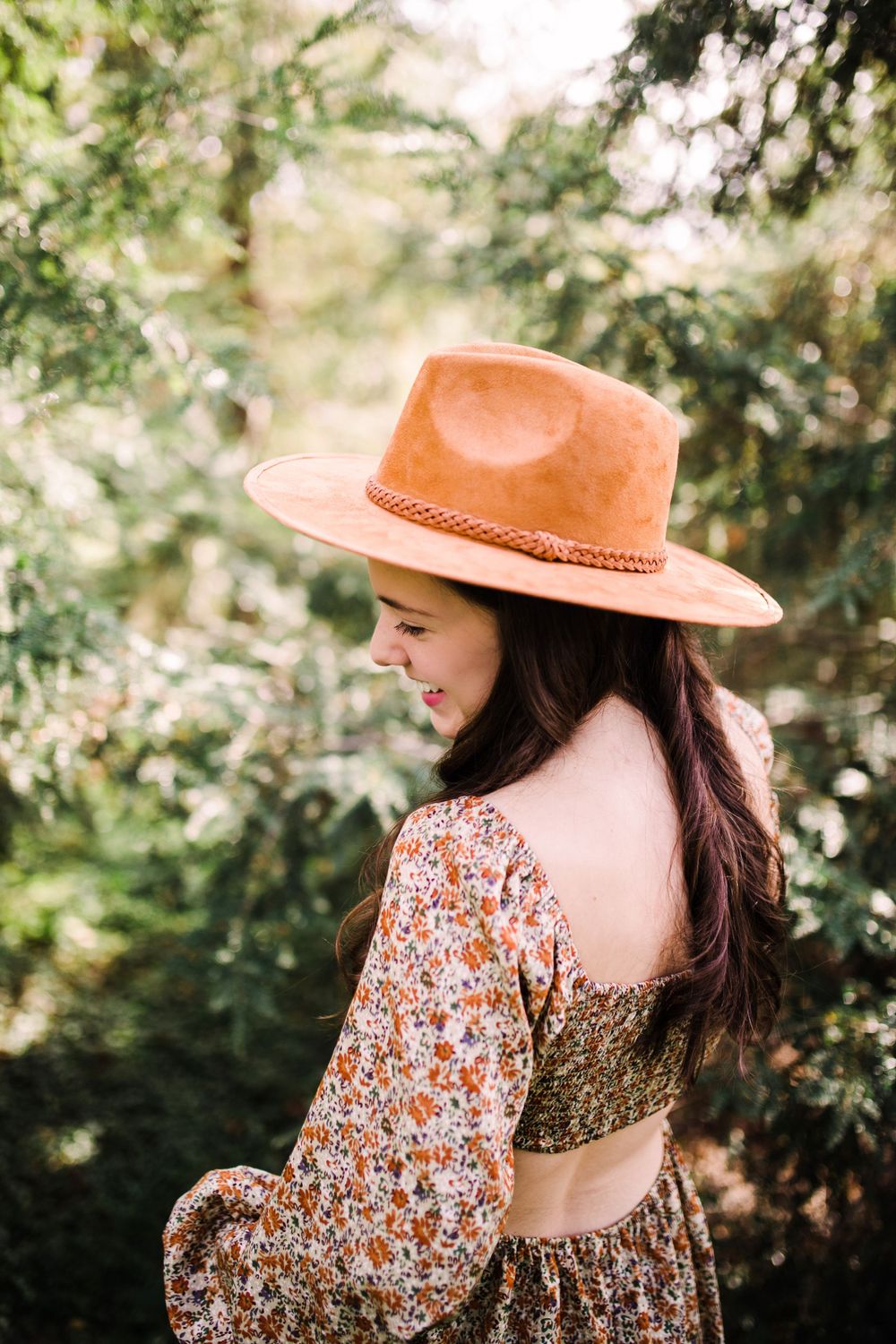 Brunette woman in floral crop top wearing a wide-brim brown hat, smiling in a lush green setting.