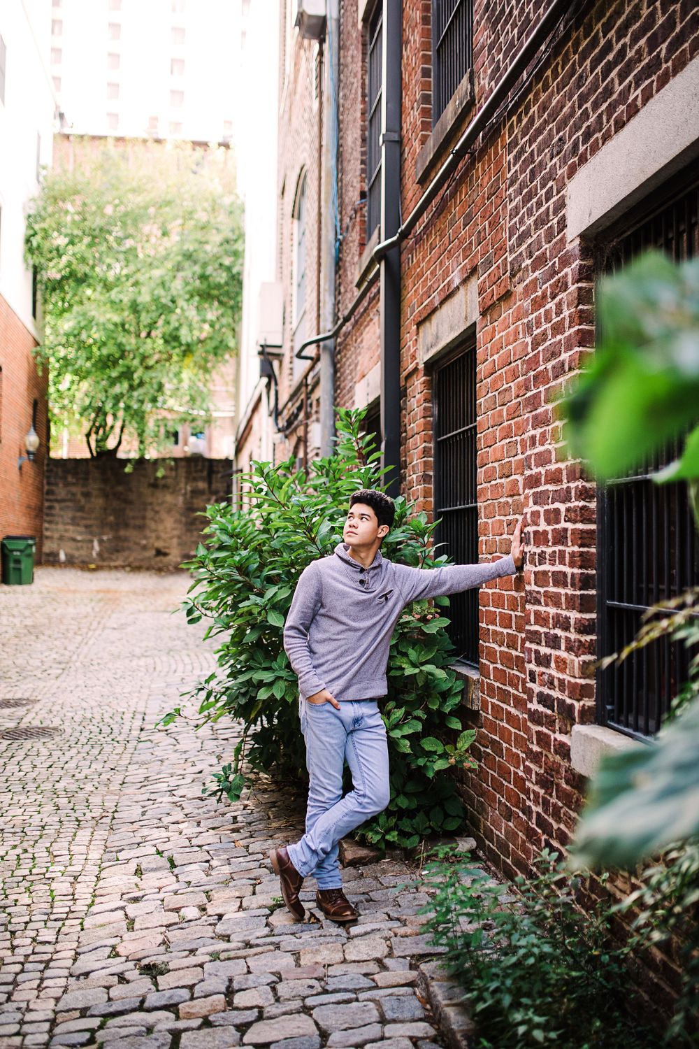 Young man leaning against brick building wall in urban alley surrounded by green foliage.