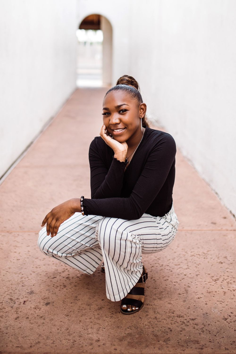 Smiling young Black woman in black top and striped pants crouches in bright white architectural hallway.