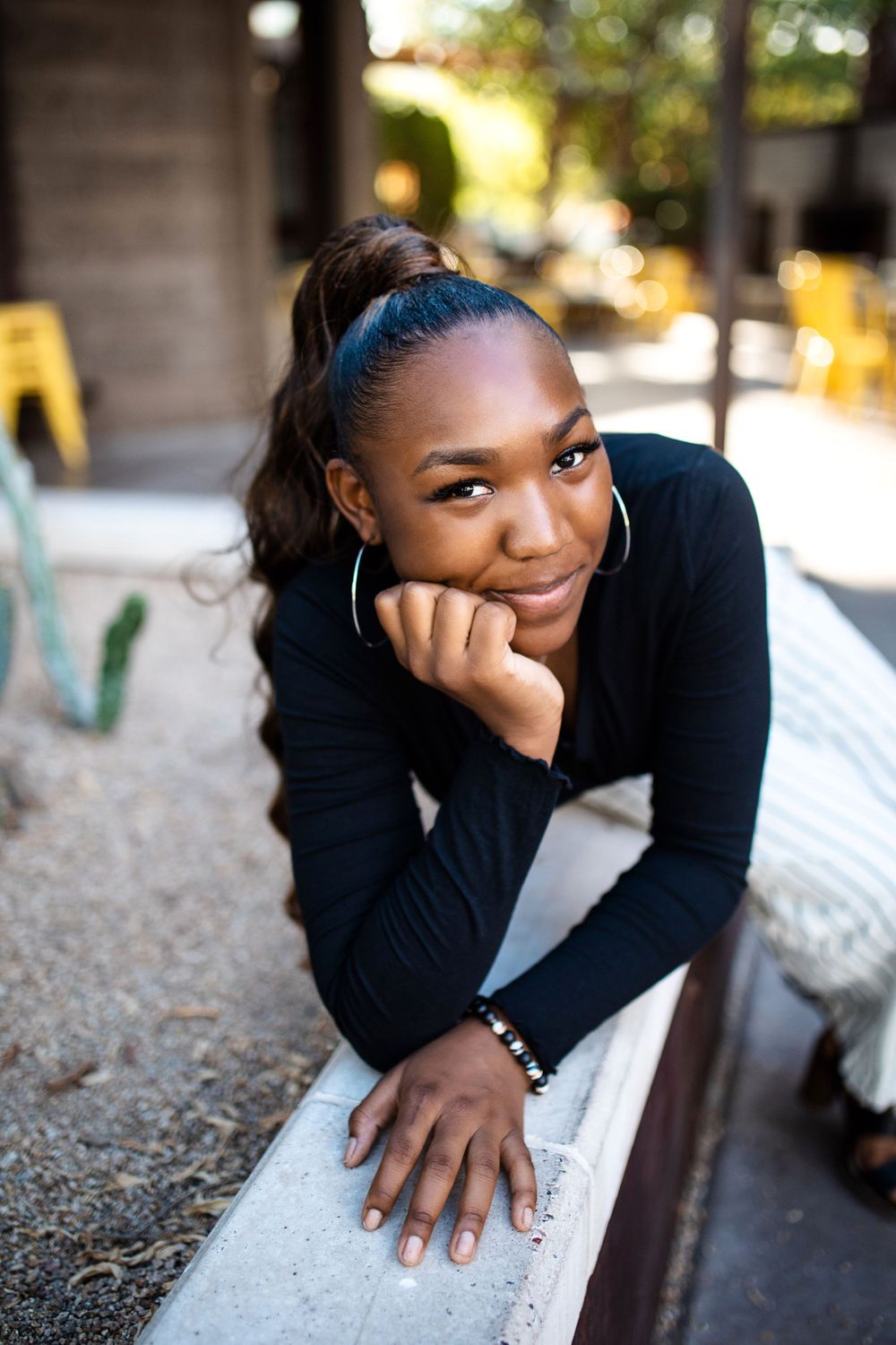 Smiling young Black woman in black long-sleeve top rests chin on hands while leaning on ledge outdoors.