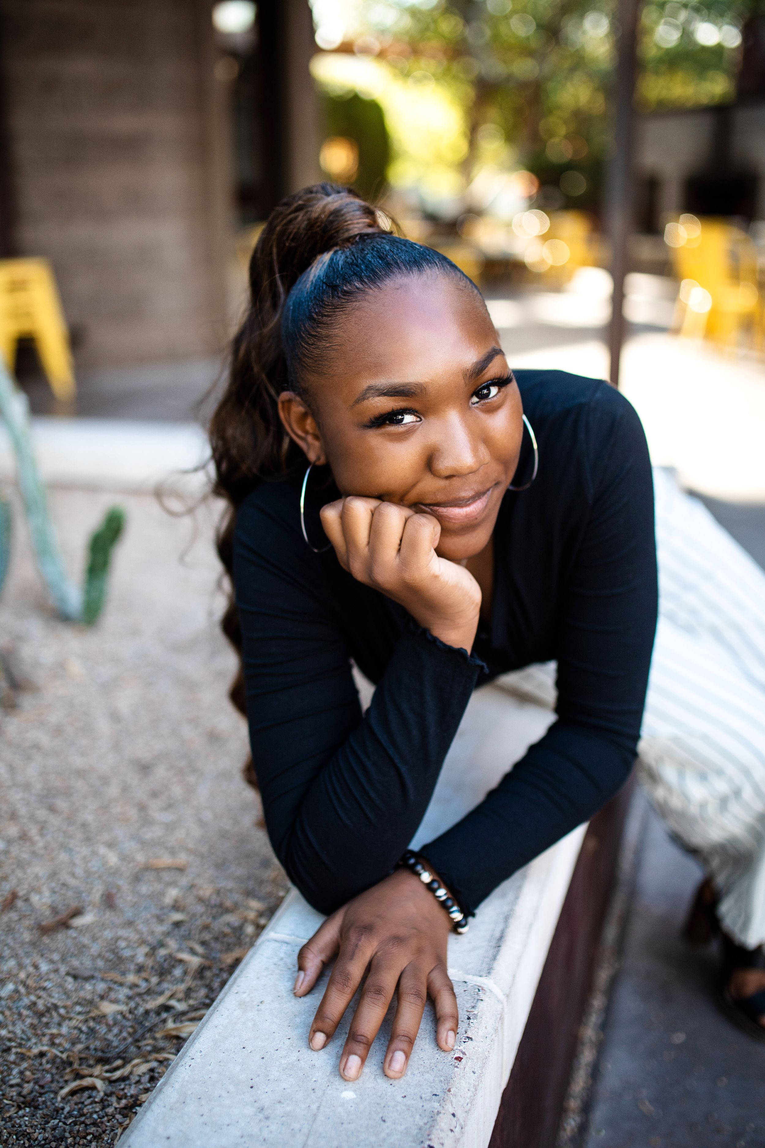 Smiling young Black woman in black long-sleeve top rests chin on hands while leaning on ledge outdoors.