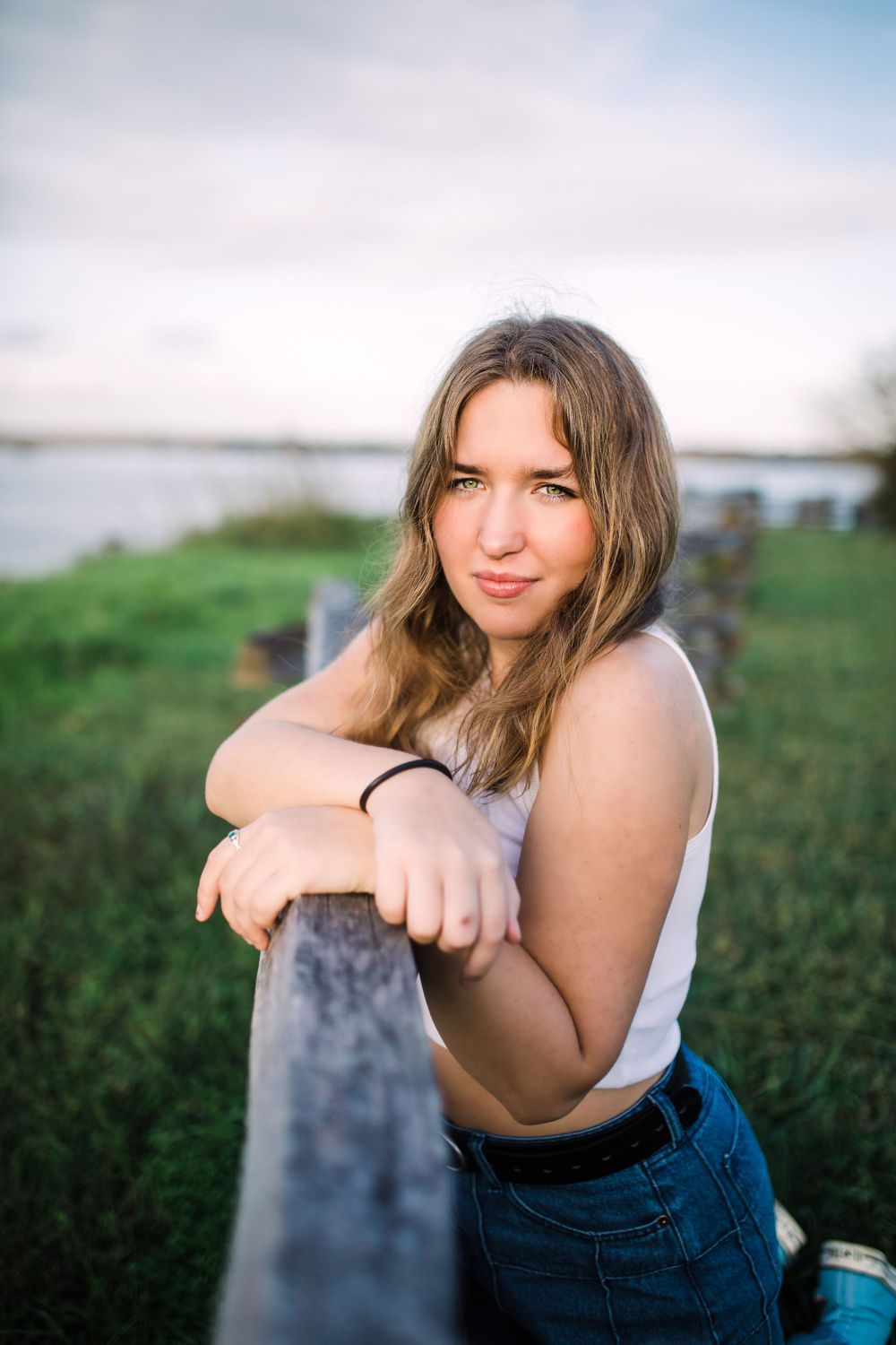 Young woman in white tank top leans on rustic wooden fence near water at sunset.