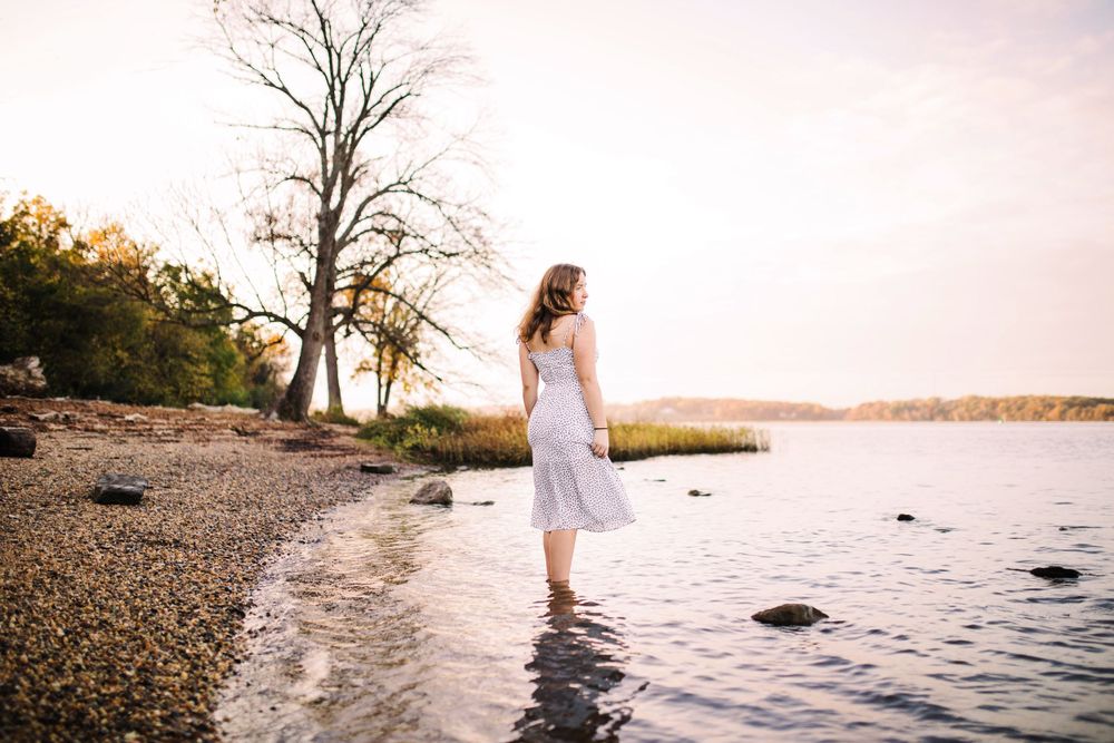Young woman in white sundress standing in shallow lake water on rocky shoreline at sunset.