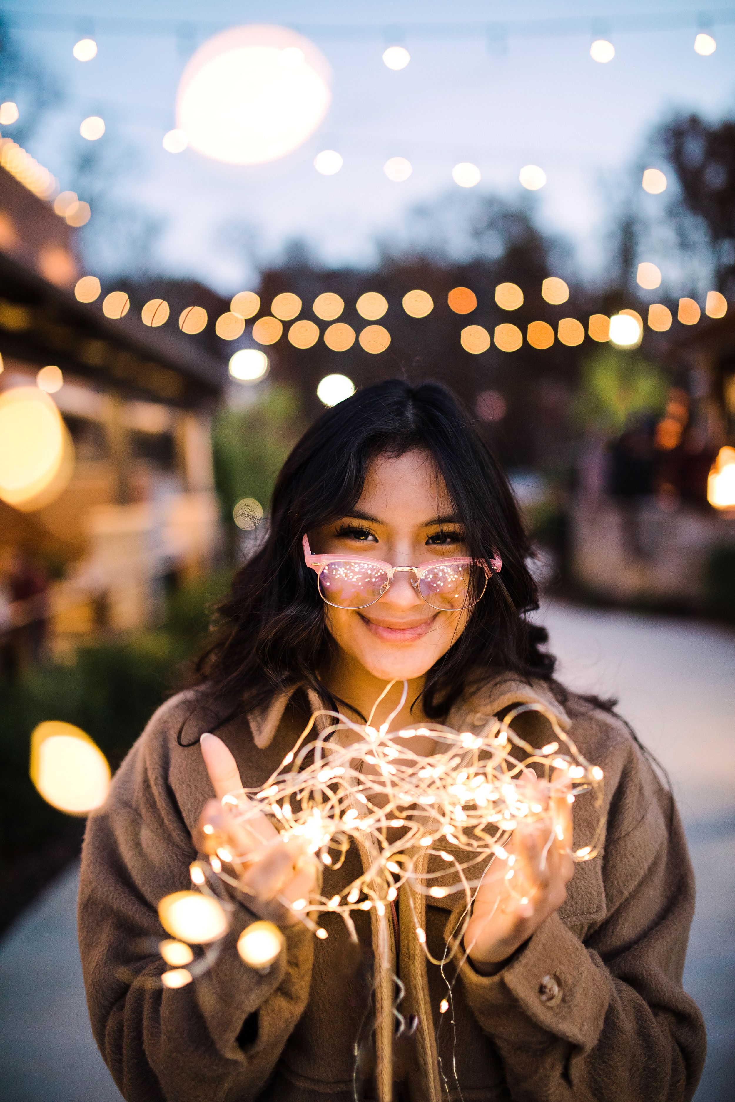 Smiling young woman with pink glasses holding glowing fairy lights under outdoor string lights at twilight.