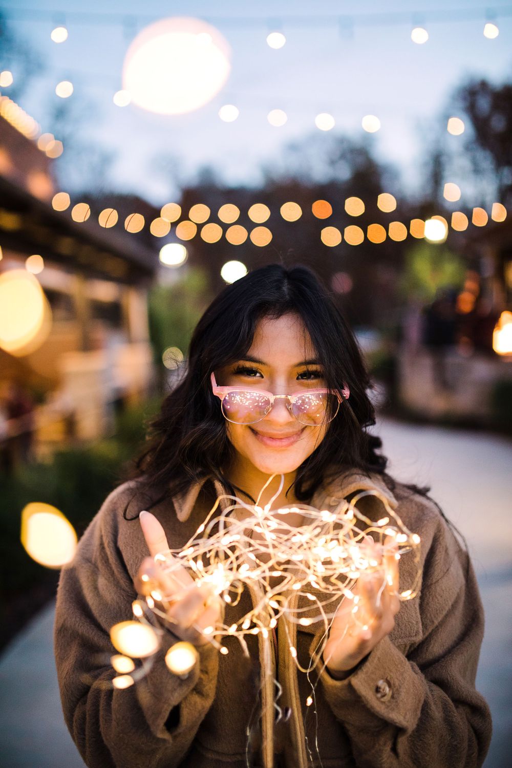 Smiling young woman with pink glasses holding glowing fairy lights under outdoor string lights at twilight.