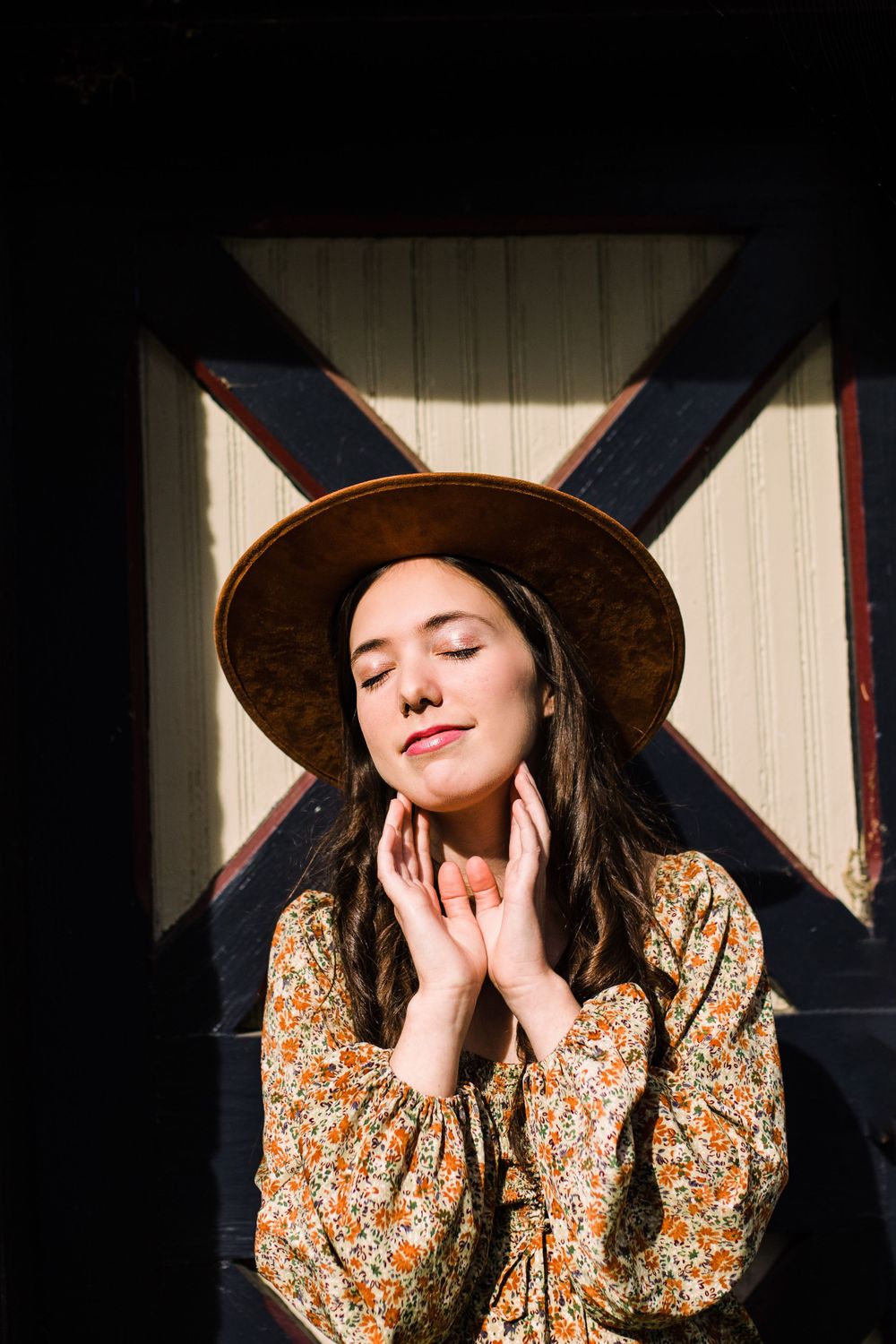 Serene young woman in floral dress and brown hat with eyes closed, hands raised to face.