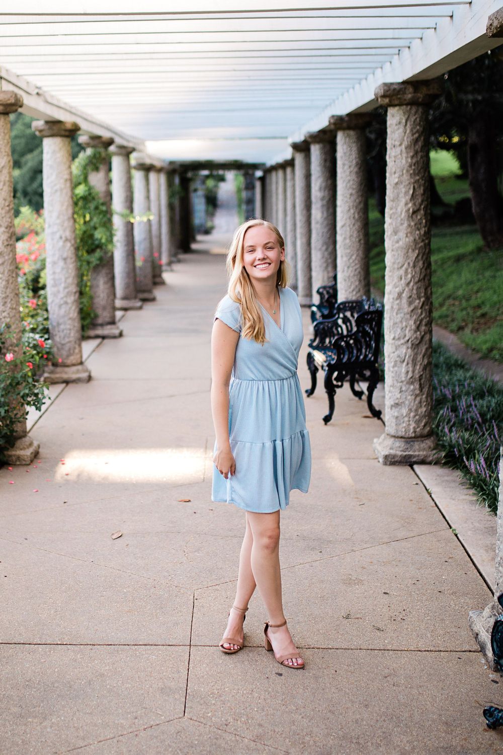 Blonde teen girl in light blue dress laughing under stone pergola with columns in park.