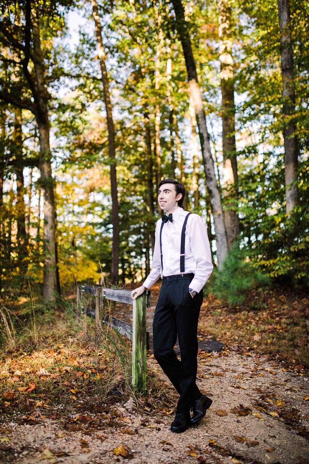 Stylish young man in suspenders and bow tie leaning on fence gazing upward in colorful autumn forest.