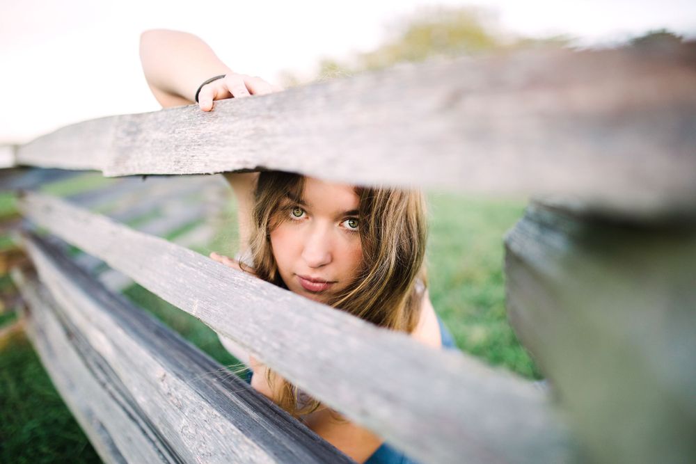 Young woman with blue eyes peeks playfully through gaps in a rustic wooden fence.