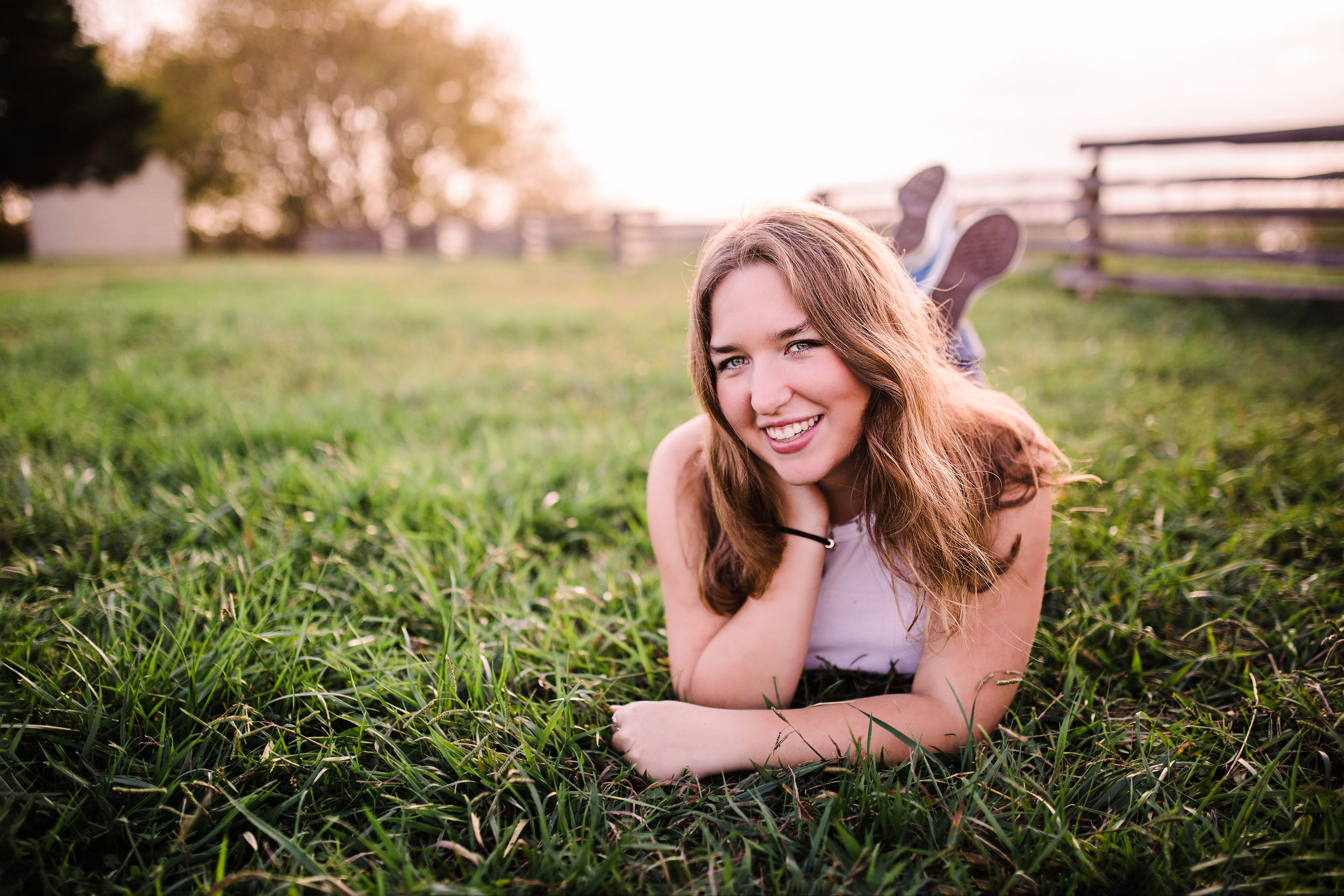Smiling young woman with long hair lies on green grass in golden sunset light.