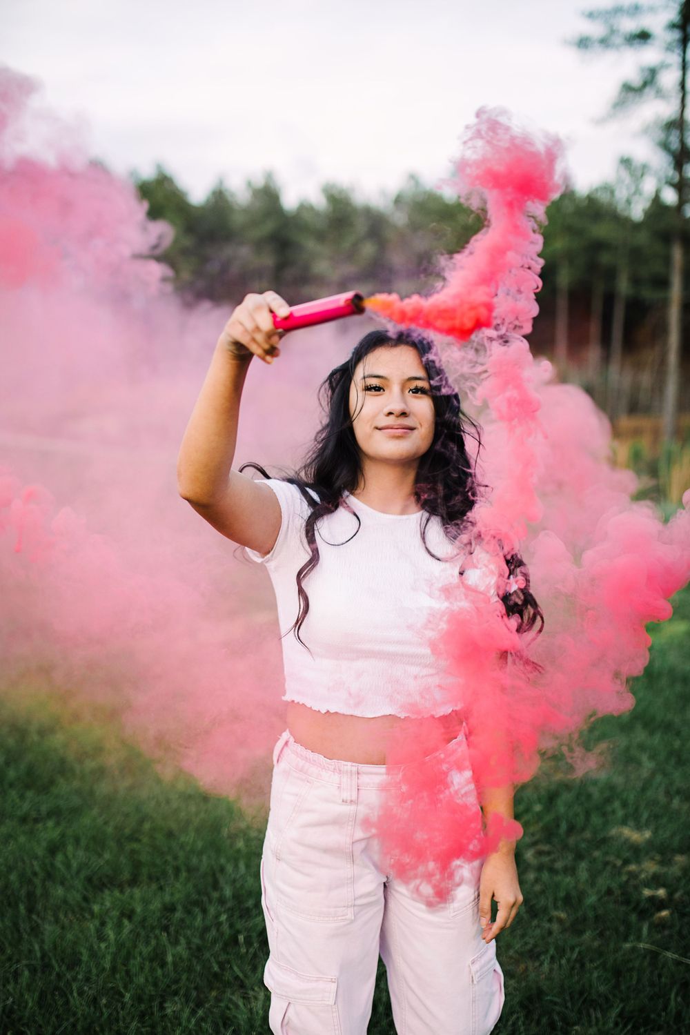 Young woman in white outfit holding pink smoke bomb in outdoor setting.