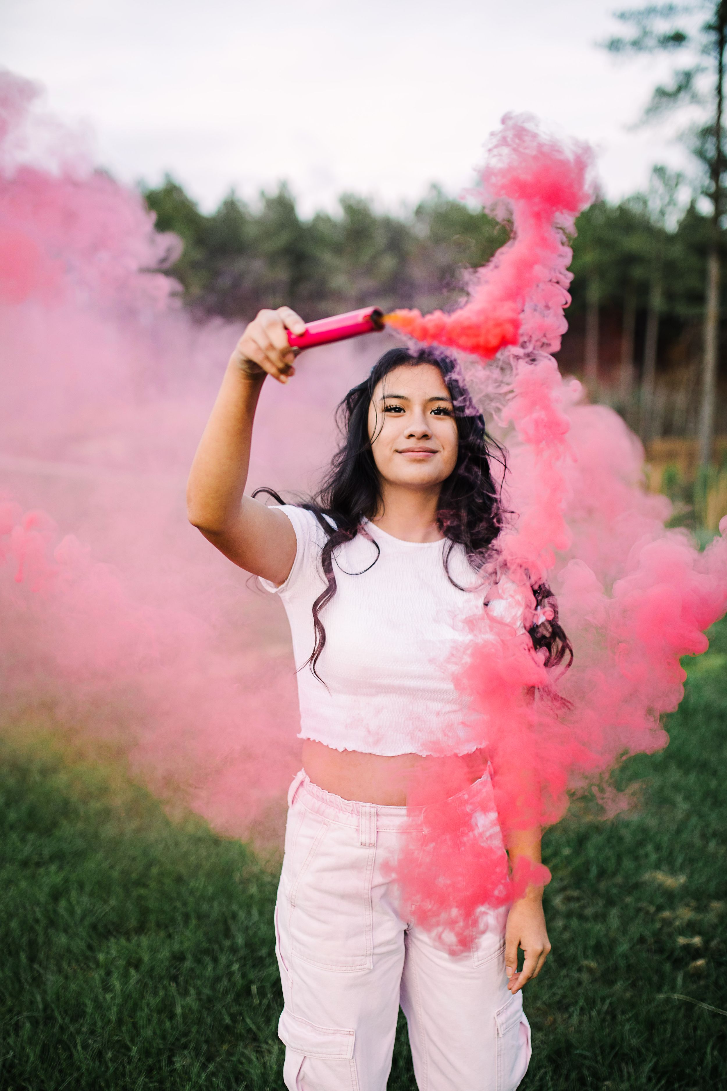 Young woman in white outfit holding pink smoke bomb in outdoor setting.