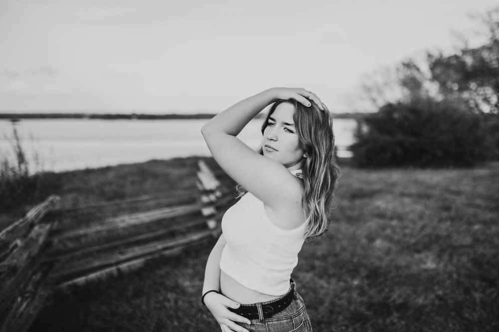 Black and white portrait of a young woman in a white tank top posing near a wooden fence outdoors.