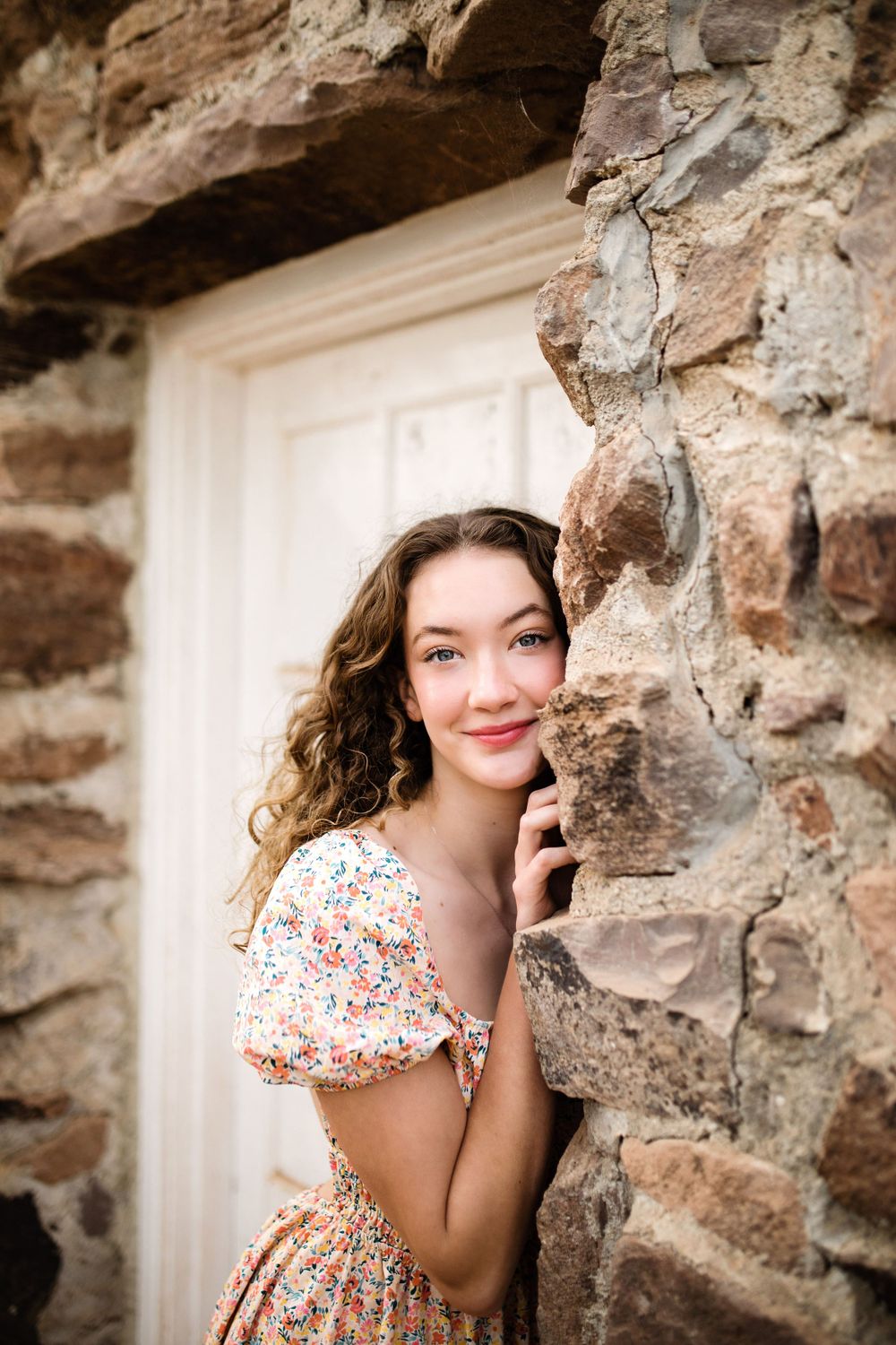 Smiling young woman with curly hair peeking around a stone wall in a floral dress.