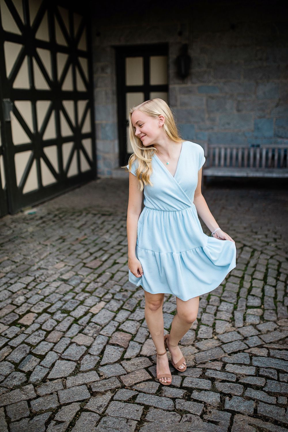 Smiling blonde teen twirling light blue dress on cobblestone courtyard near timber-frame building.