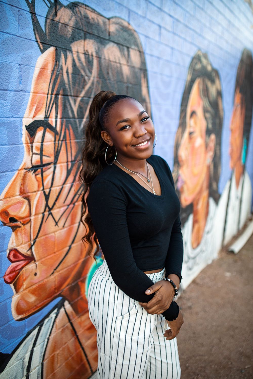 Smiling young Black woman in black top and striped pants poses in front of colorful urban mural.