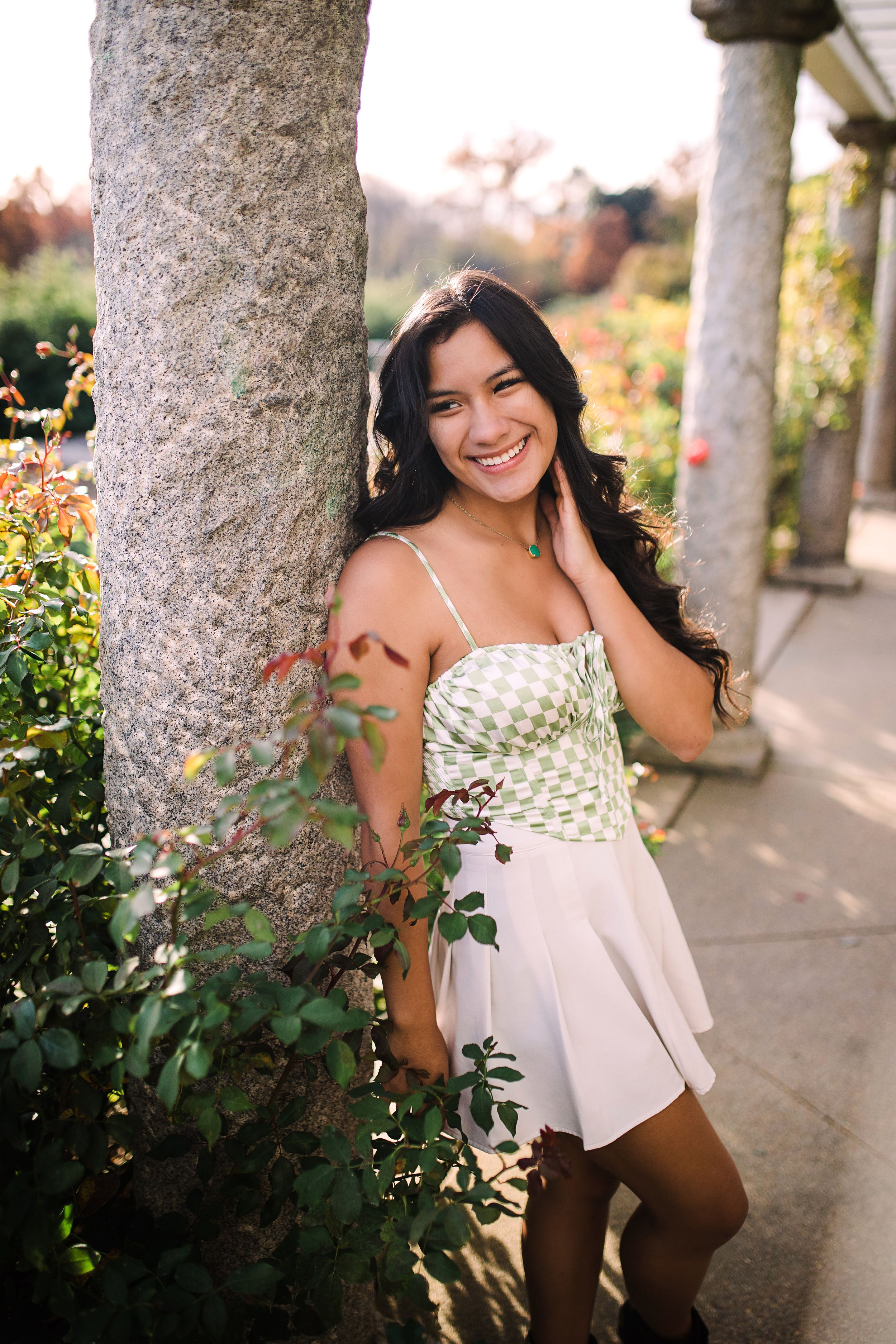 Smiling young woman in green and white dress leans against a stone column in a blooming garden.