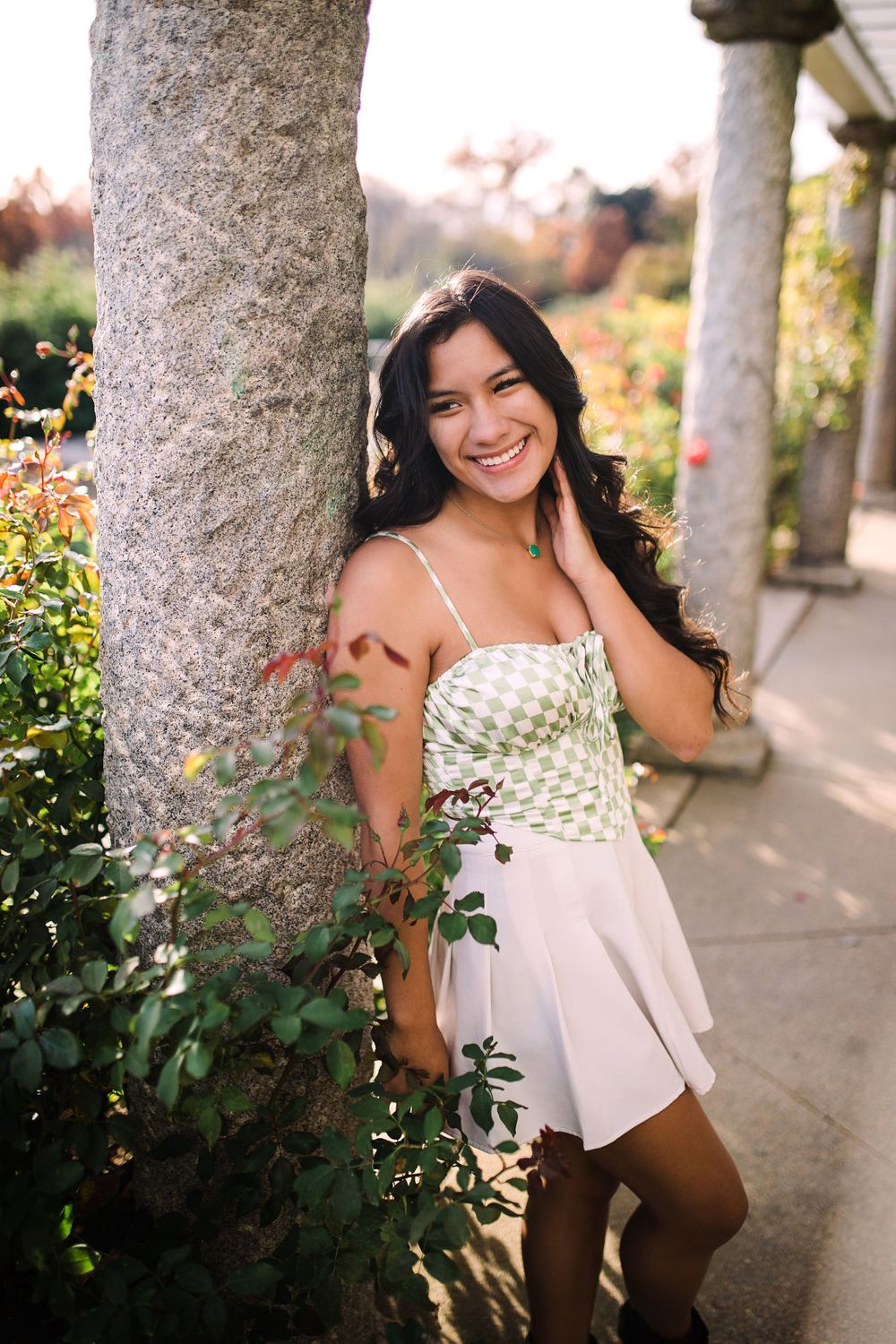 Smiling young woman in green and white dress leans against a stone column in a blooming garden.