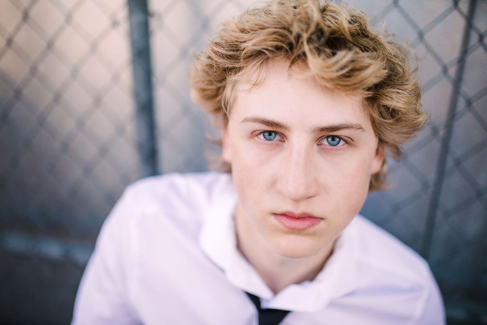 Close-up portrait of blue-eyed young man with curly blonde hair in white shirt near chain-link fence.