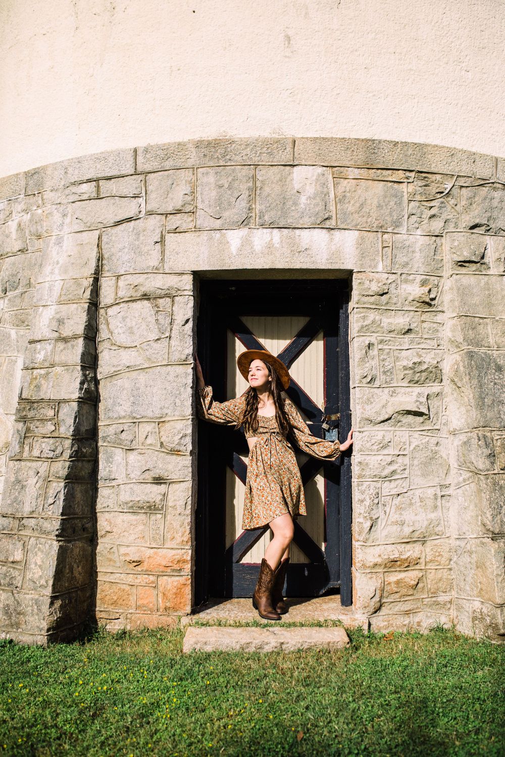 Young woman in floral dress and cowboy boots posing in stone archway doorway in sunlight.