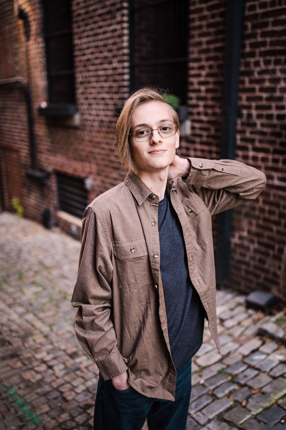 Young man with blonde hair and glasses posing casually in brown jacket on urban cobblestone street.