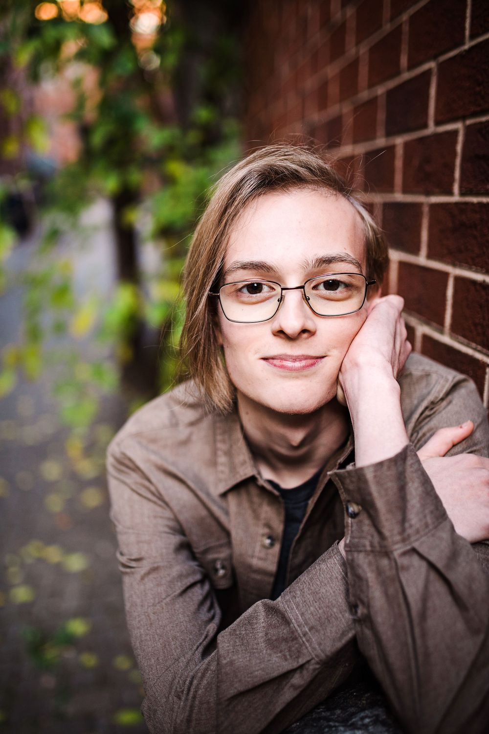 Close-up portrait of young person with glasses resting head on hand against red brick wall.
