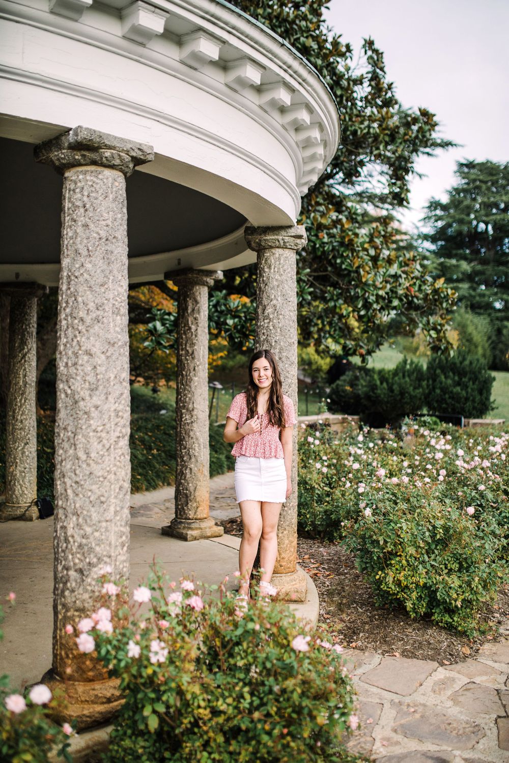 Young woman in pink top and white skirt posing between granite columns of elegant garden pavilion.