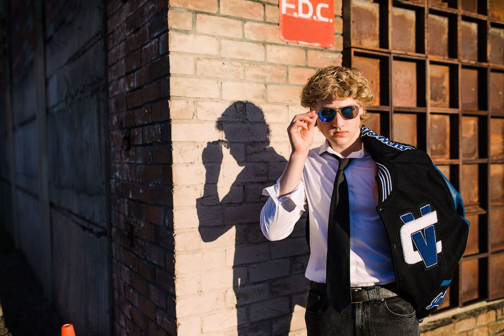 Young man in varsity jacket adjusting sunglasses beside brick wall with bold shadow in sunlight.