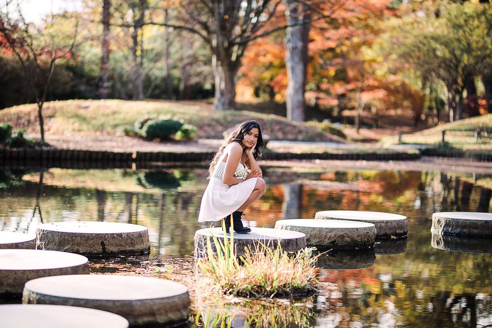 Bride in white gown poses gracefully on stepping stones over a serene pond in an autumn Japanese garden.
