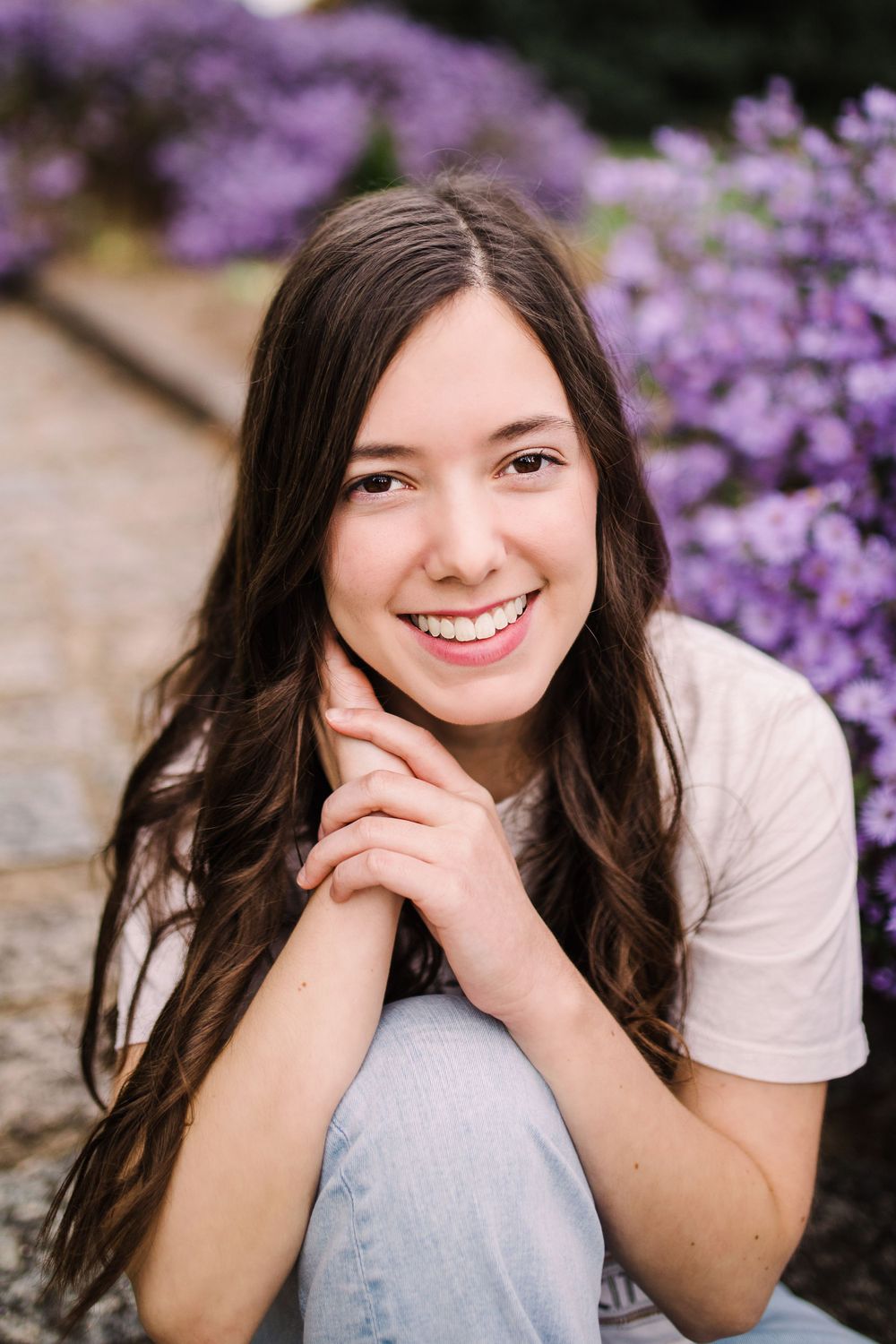 Smiling teenage girl with dark hair posing with hands clasped near purple flowering bushes outdoors.