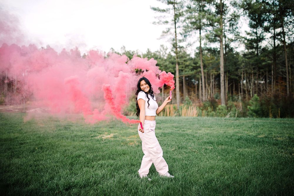 Young woman in white outfit twirling with vibrant pink smoke bombs in an open grassy field.