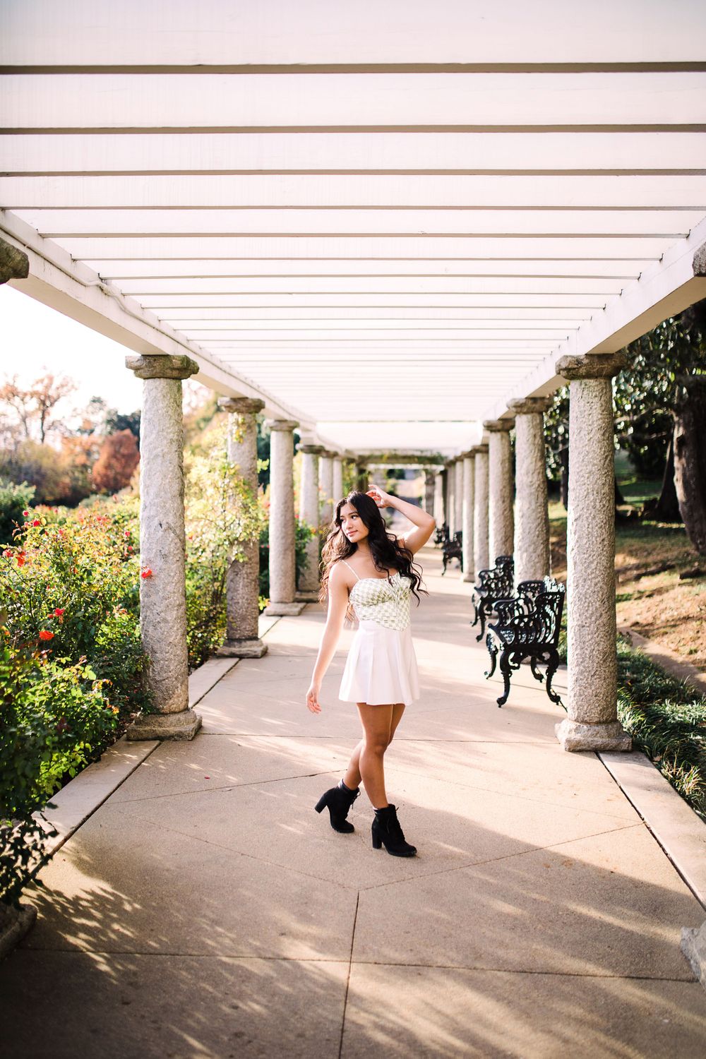 Young woman in white dress and black boots walks gracefully through a pergola-covered garden pathway.
