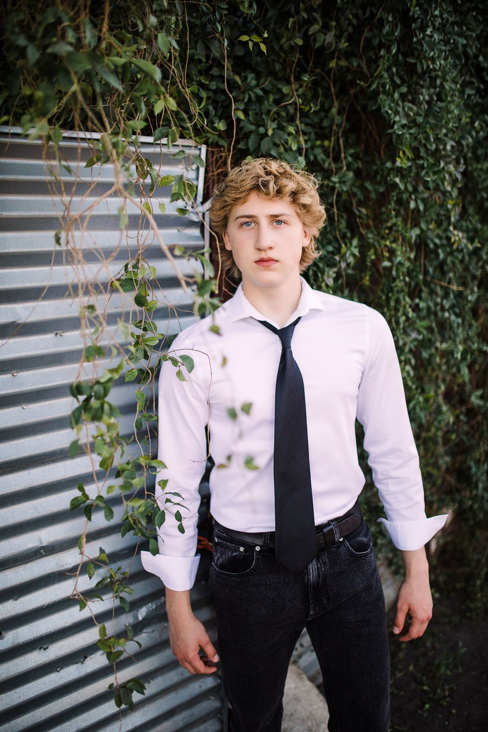 Young man in white shirt and black tie standing before corrugated metal wall with green ivy.