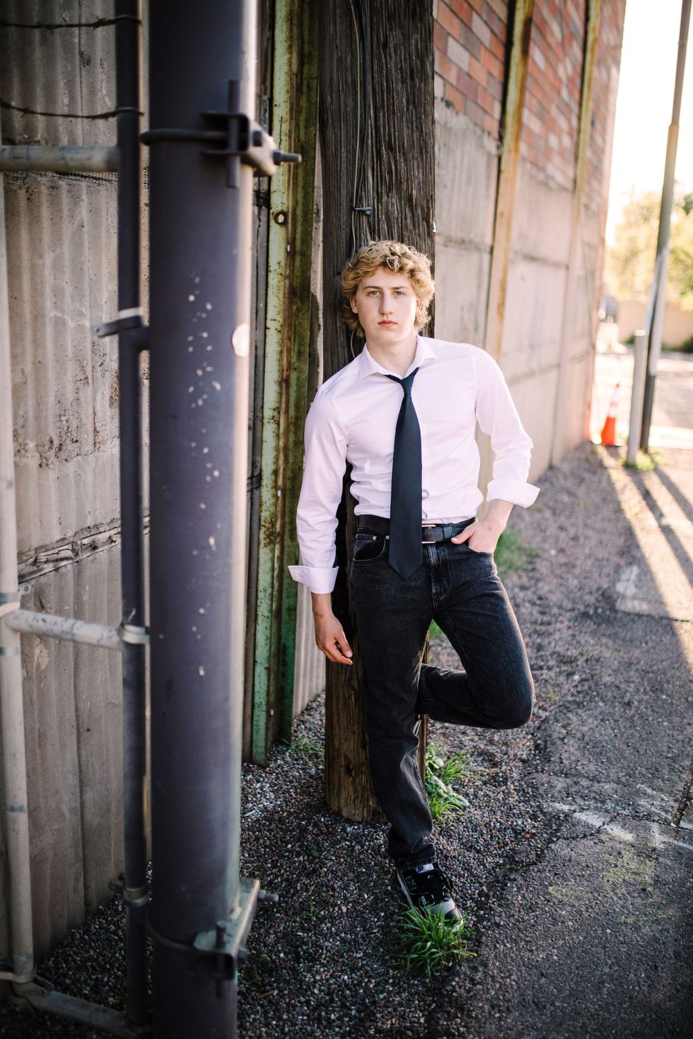Teen boy in white shirt and black tie leaning against pole in urban alley with brick wall.
