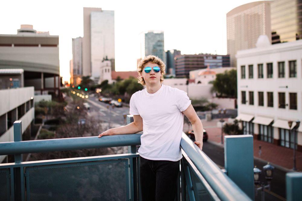 Young man in white tee and blue sunglasses poses on urban rooftop balcony with city skyline at sunset.