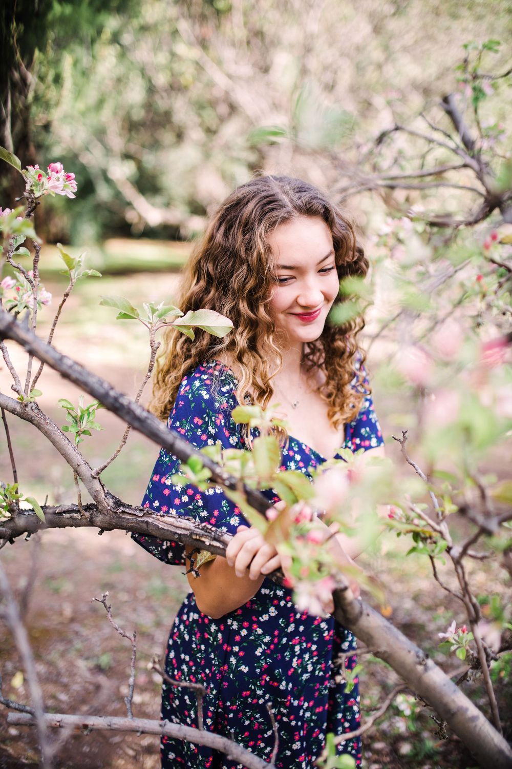 Young woman in navy floral dress smiling among blooming spring tree branches.