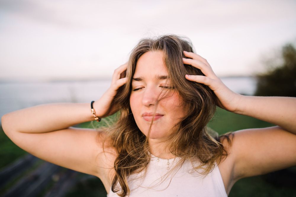 Woman with wavy brown hair holds head with both hands outdoors near water at dusk.