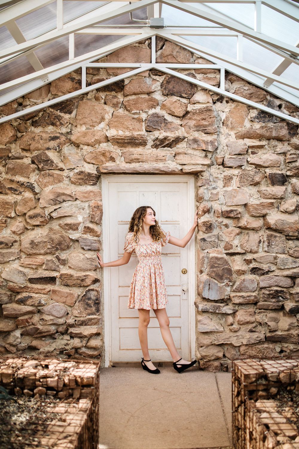 Young woman in floral dress posing by white door against rustic stone greenhouse wall.