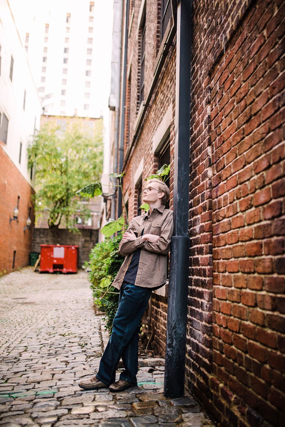 Young man in brown jacket leaning against brick wall on cobblestone alley looking upward thoughtfully.