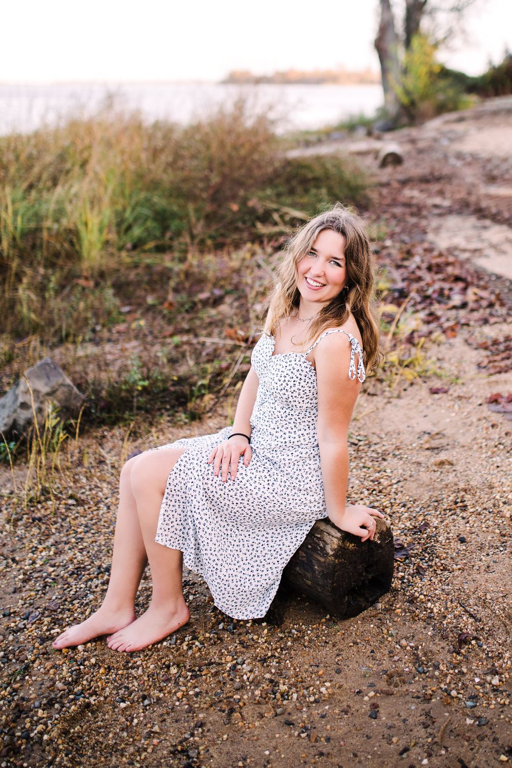 Happy young woman in white spotted sundress sitting barefoot on log along scenic shoreline.