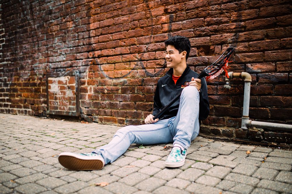 Smiling young man sitting against brick wall on cobblestone street with lacrosse stick nearby.
