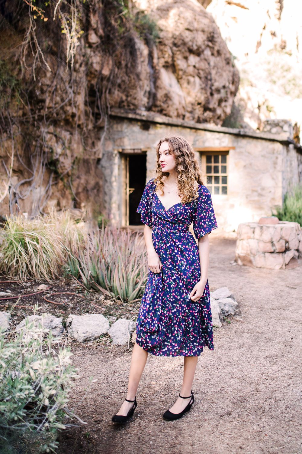 Young woman in navy floral midi dress posing outdoors near a rocky hillside and stone building.