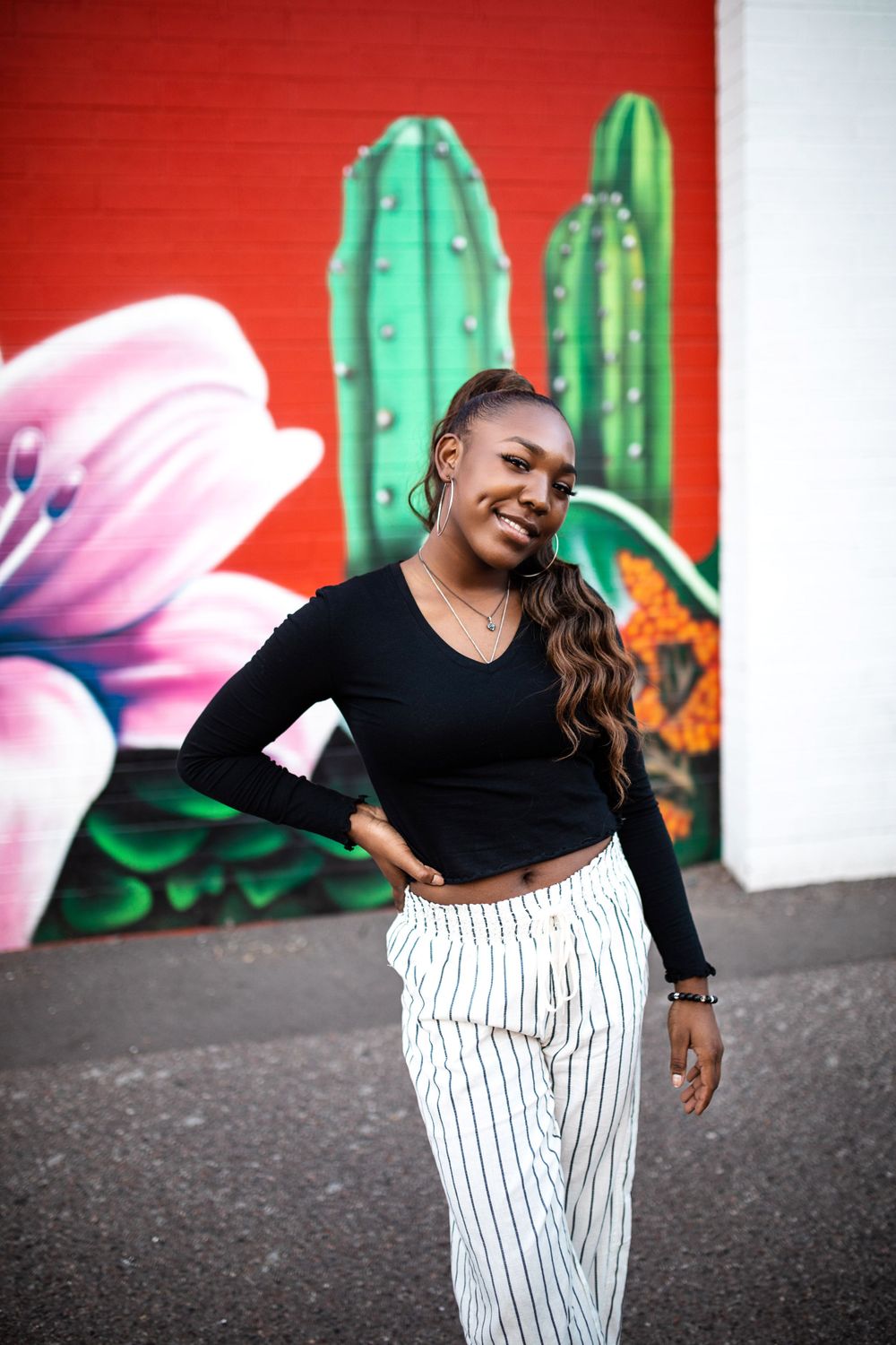 Smiling young Black woman in black crop top and striped pants poses before vibrant cactus mural.