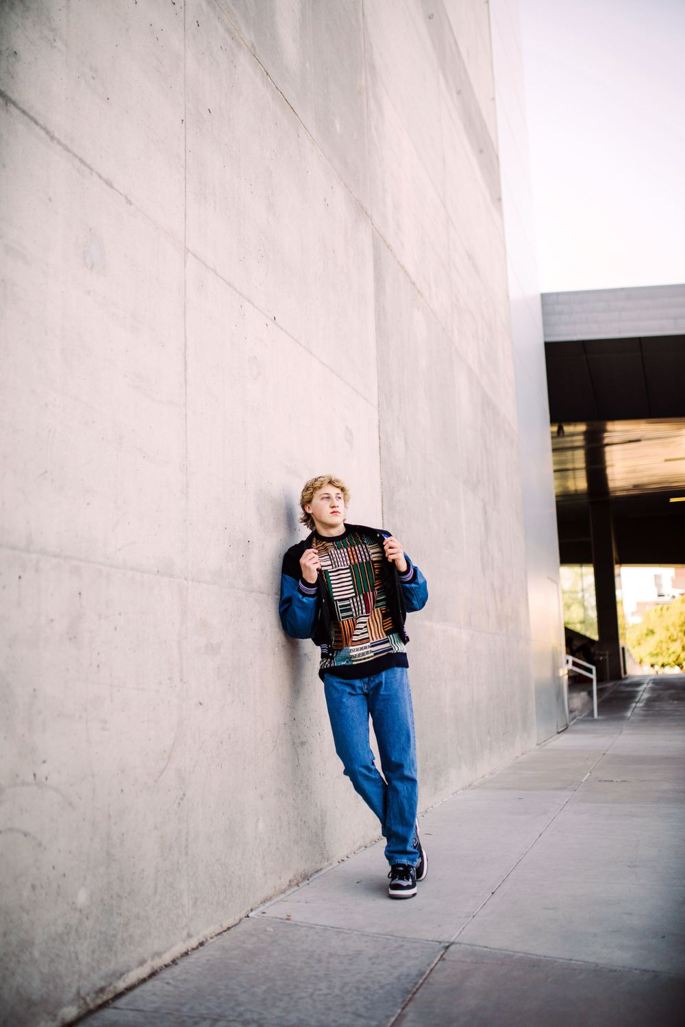Teen boy in varsity jacket leaning against concrete wall in jeans outdoors.
