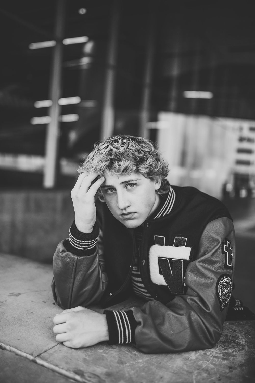 Black and white portrait of curly-haired teen in varsity jacket resting on concrete ledge.