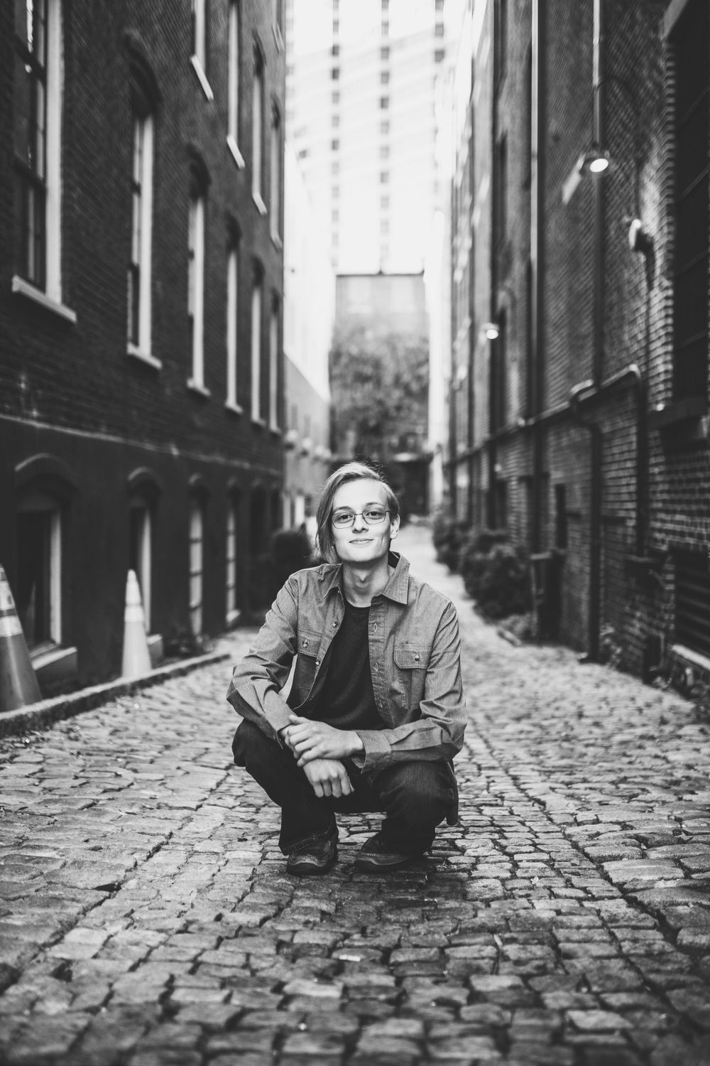 Black and white portrait of young man with glasses crouching on historic cobblestone alley.