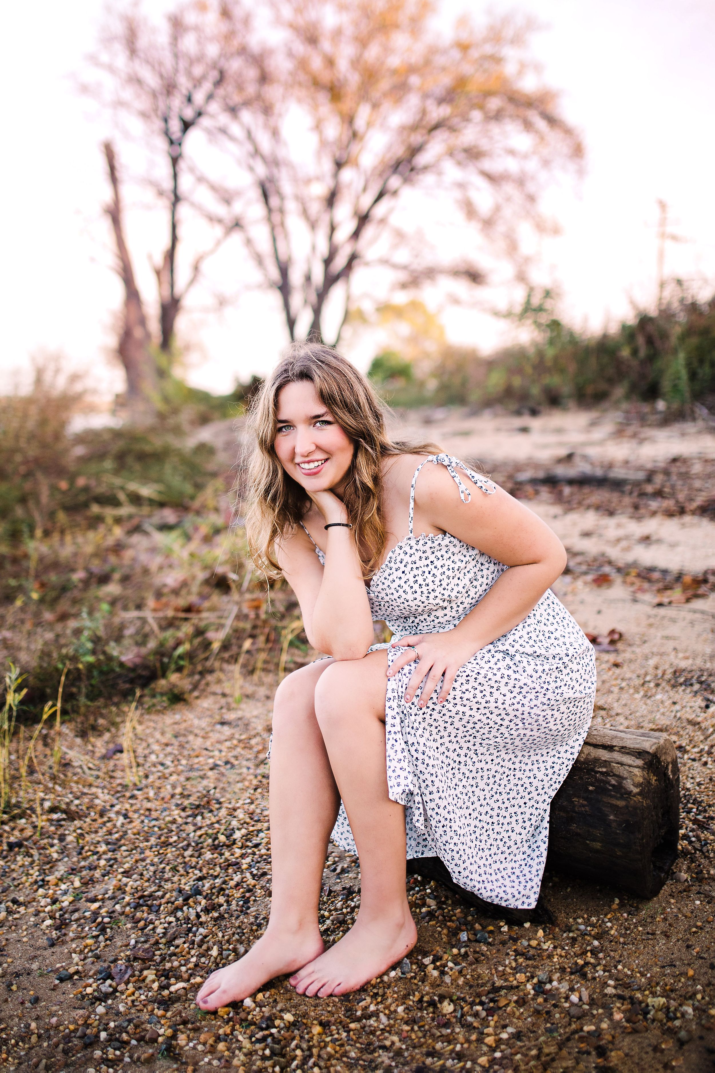 Smiling young woman in white floral dress sitting barefoot on log on rocky beach at sunset.